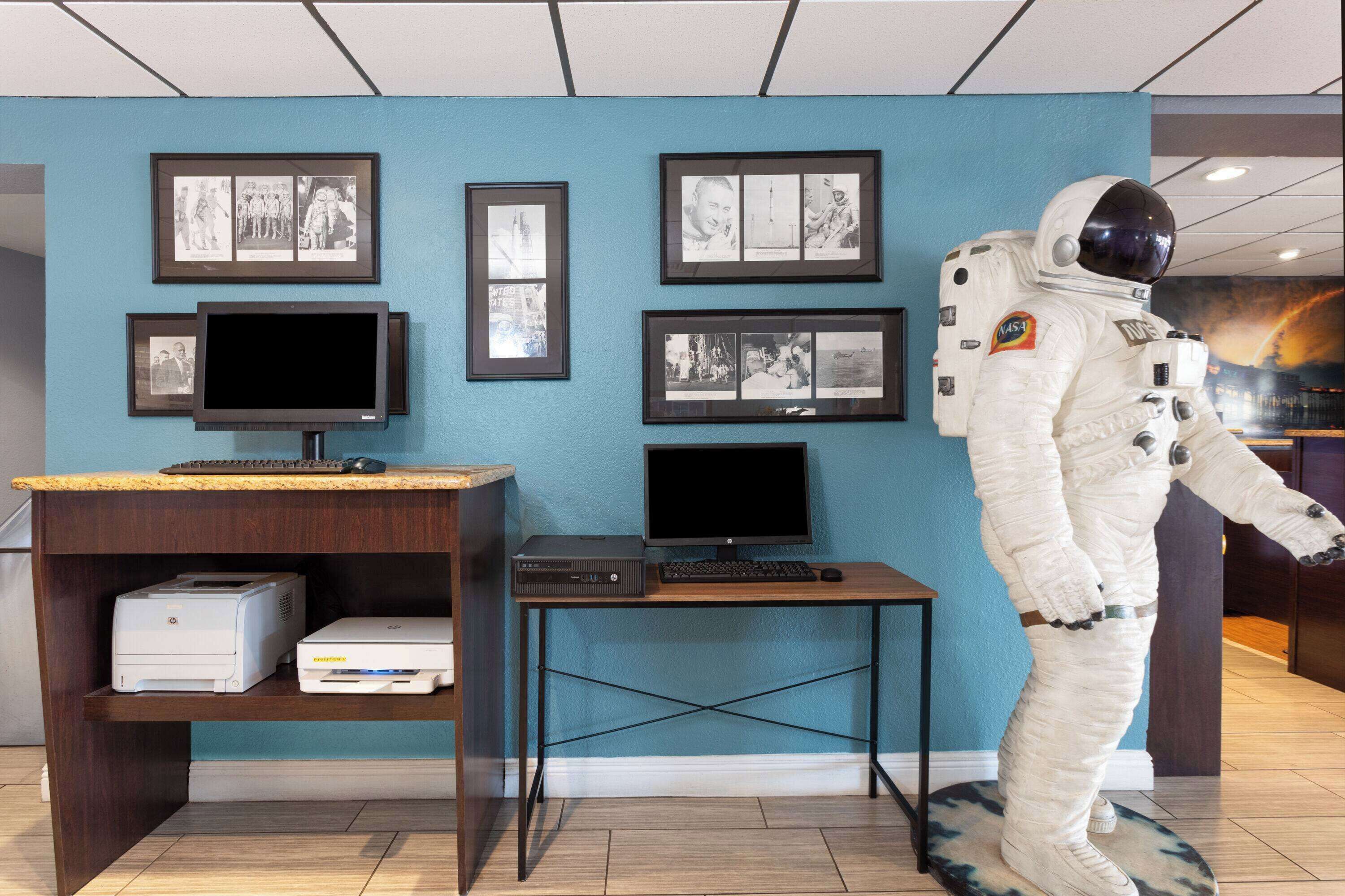 Une pièce comprenant un bureau, deux écrans d’ordinateur, une imprimante et une figurine de combinaison spatiale.