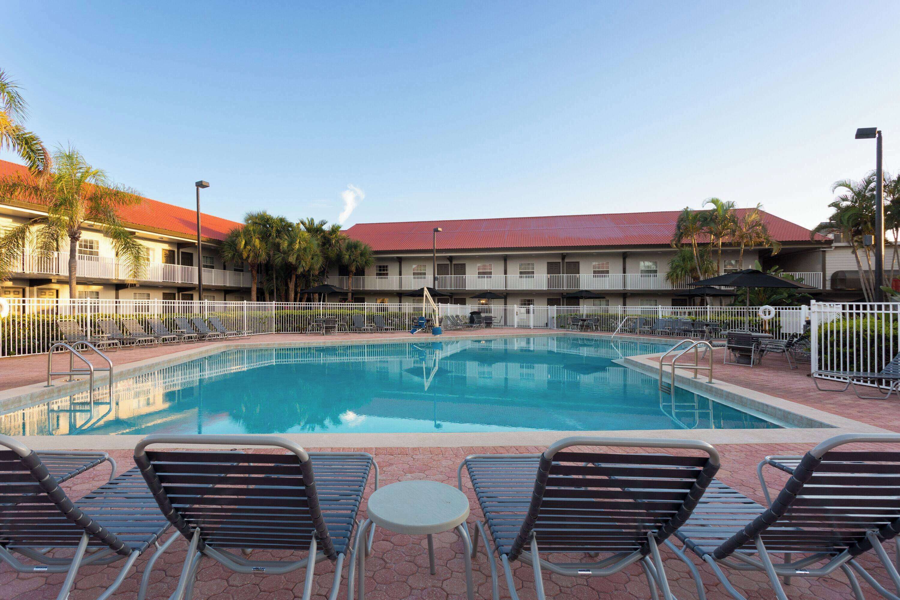 A swimming pool with lounge chairs and umbrellas, surrounded by a fence and palm trees.