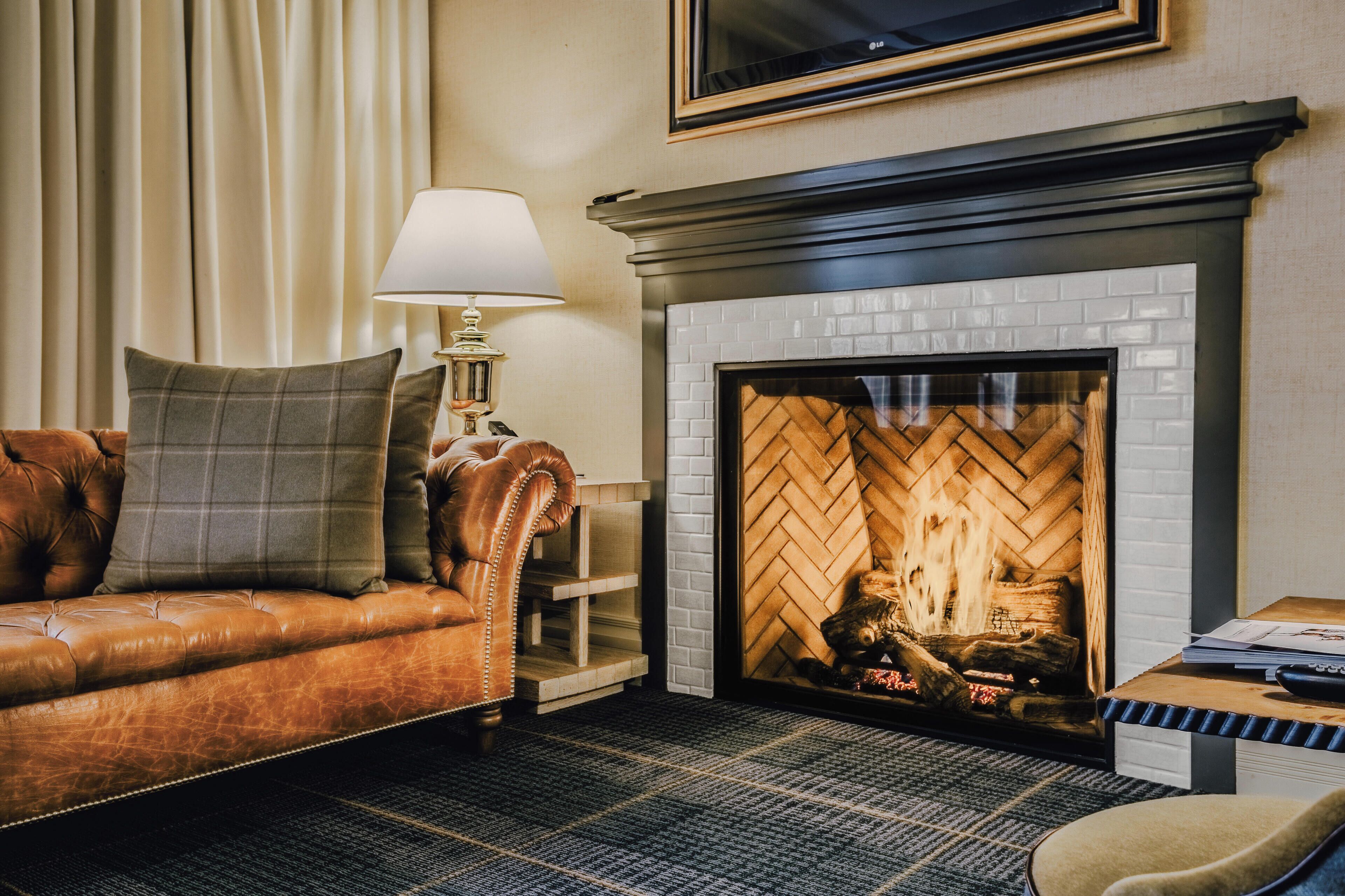 A cozy living room with a leather sofa, a side table with a lamp, and a fireplace with a brick surround.