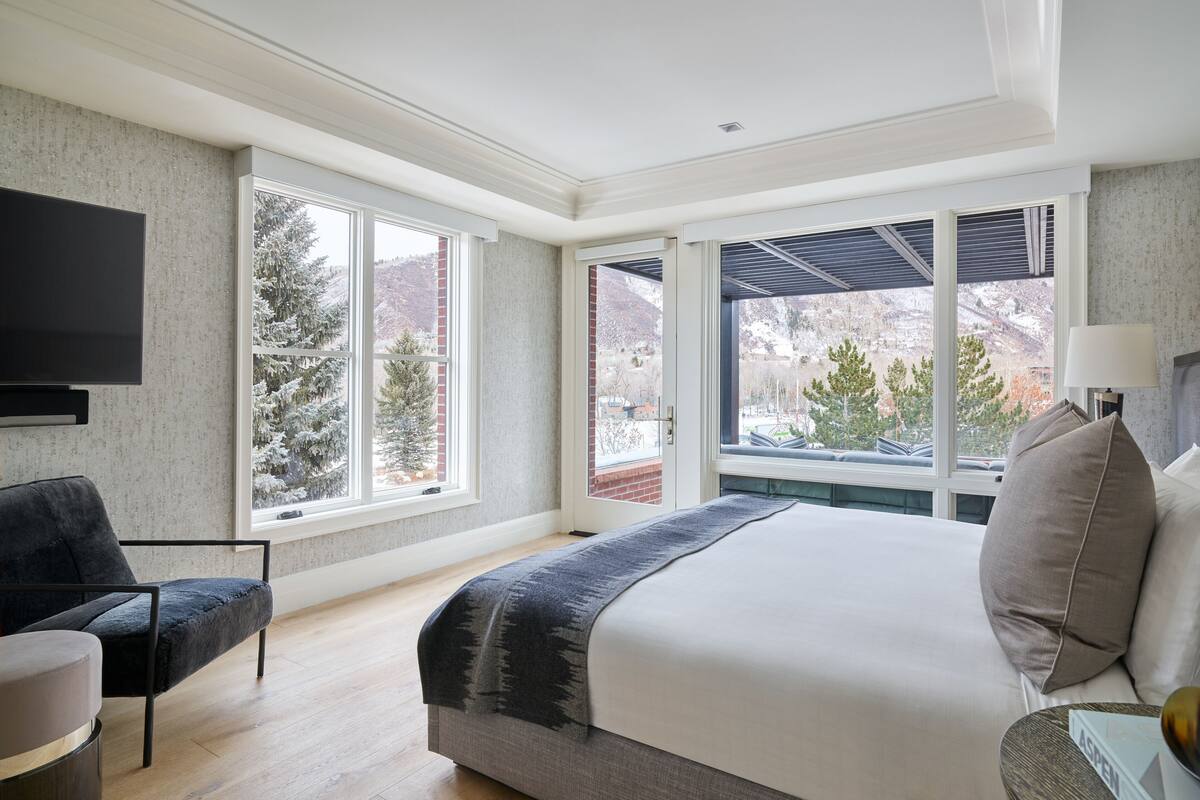 Une chambre d’hôtel moderne dotée d’un grand lit, d’un téléviseur à écran plat, d’un coin salon avec un fauteuil et de grandes fenêtres offrant une vue sur des arbres et des montagnes enneigés.