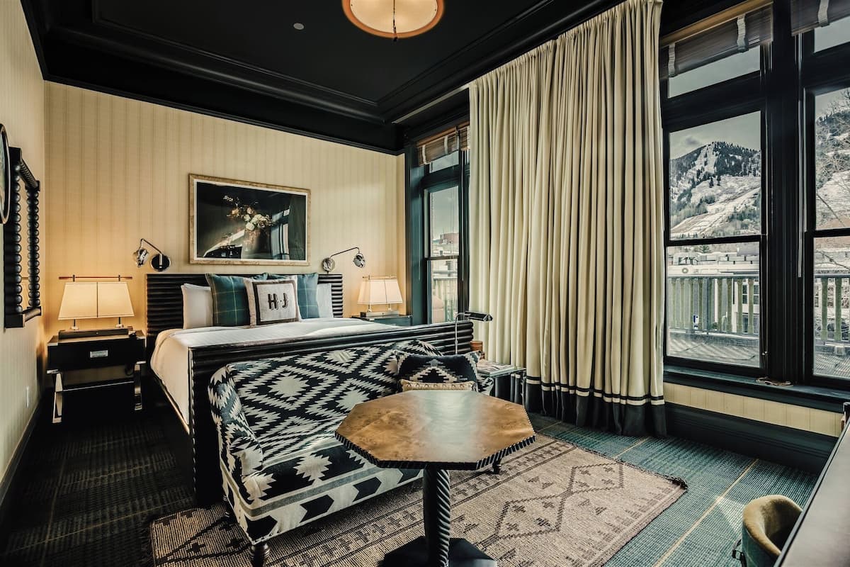 Une chambre d’hôtel avec un lit, une petite table, une fenêtre avec des rideaux, un tableau au mur et une vue sur un paysage enneigé.