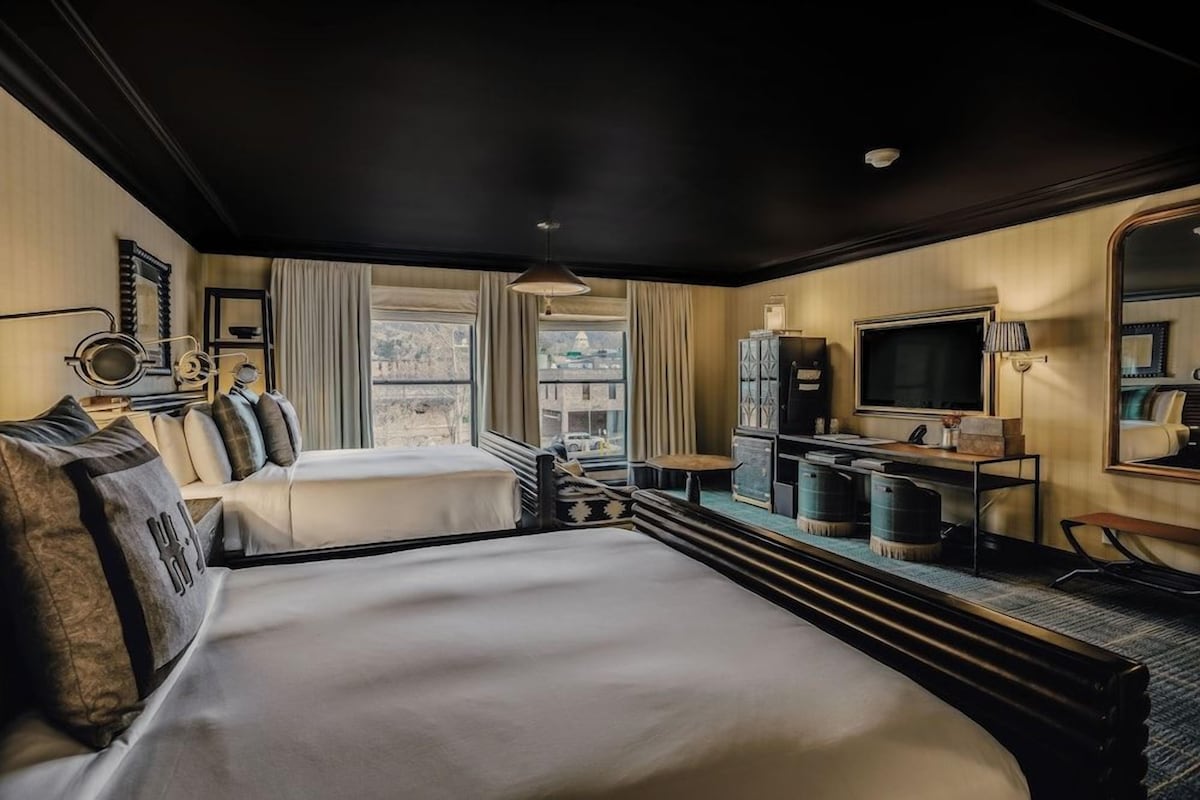 A hotel room with two beds, a television, a desk, and a view of a building through the window.
