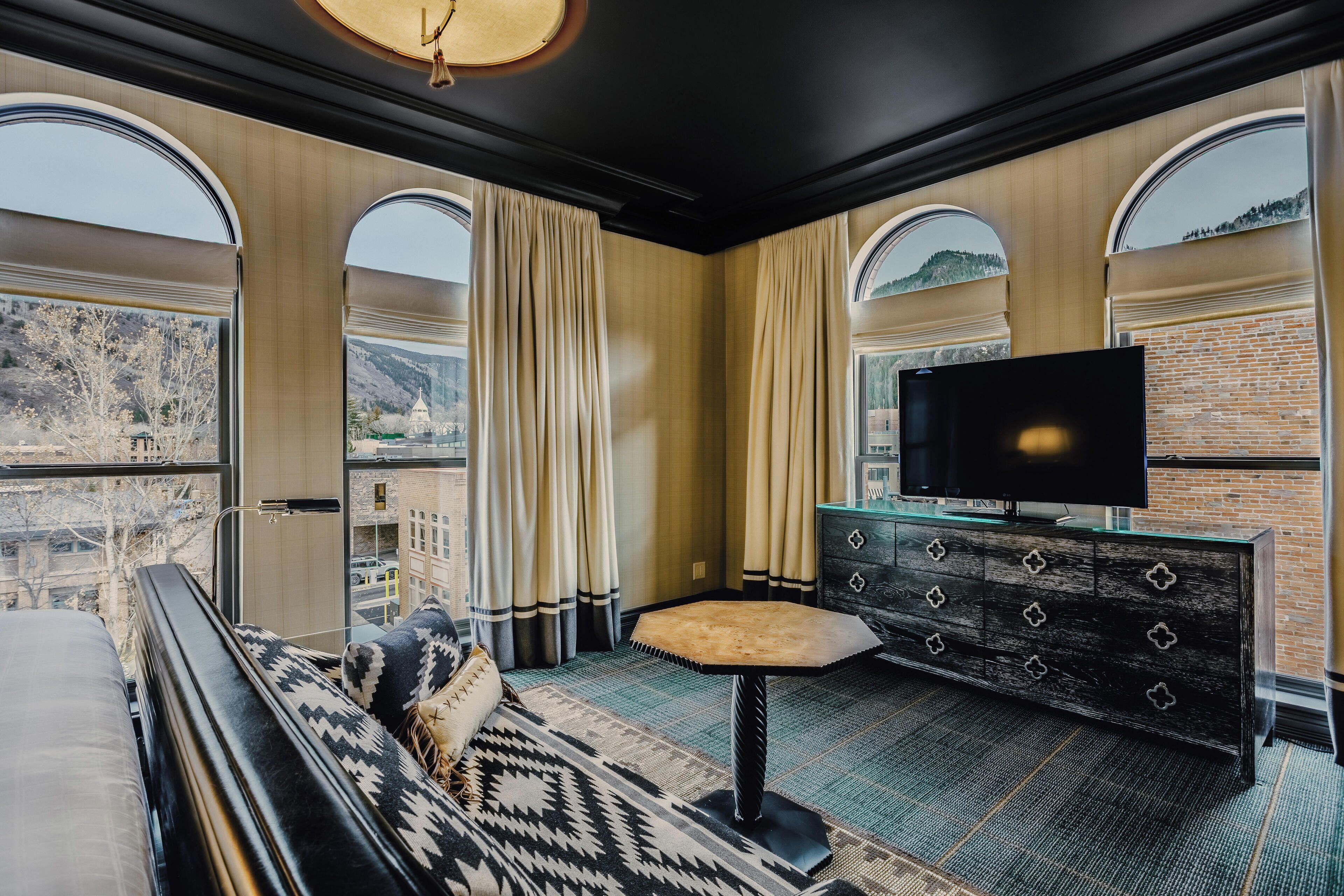 A modern hotel room with a large bed, a TV, a small table, and a view of the city through large windows.