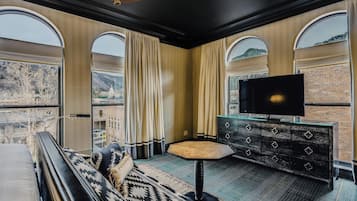 A modern hotel room with a large bed, a TV, a small table, and a view of the city through large windows.