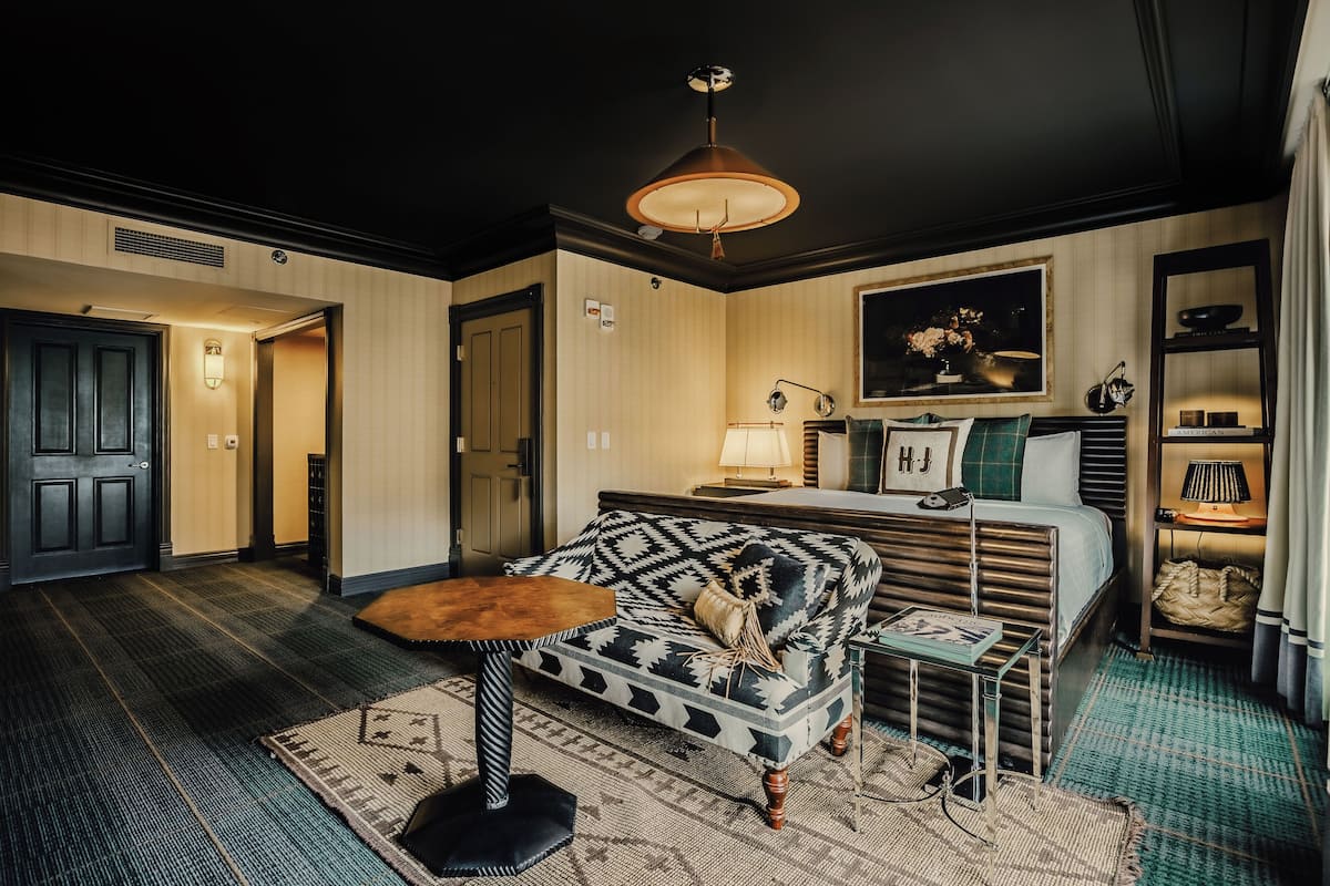 Une chambre d’hôtel avec un lit, un canapé, une petite table et une lampe. Il y a un tableau au mur et une étagère avec des objets décoratifs.