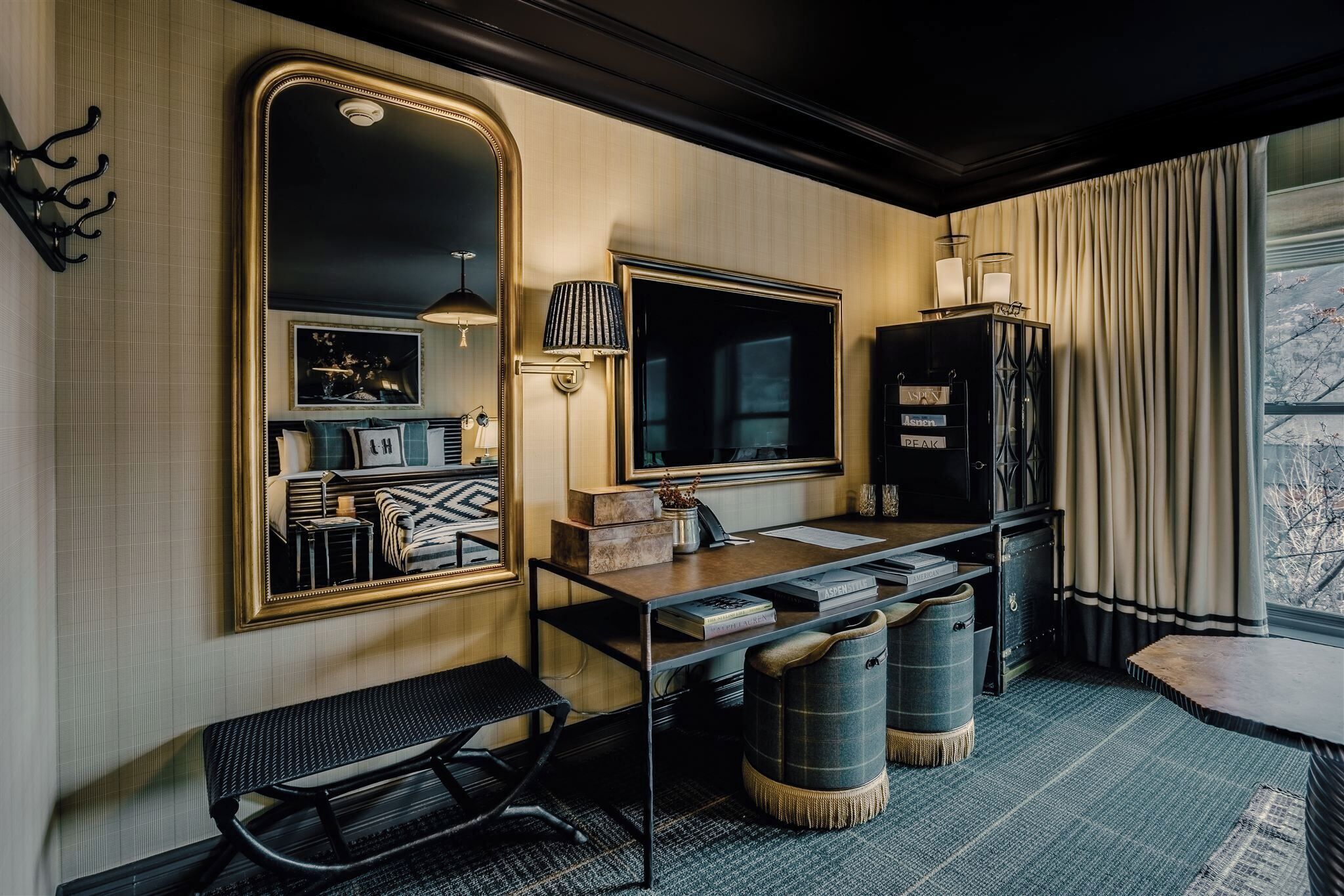 A hotel room with a large mirror, a desk with a lamp, a bench, and a view of trees through the window.