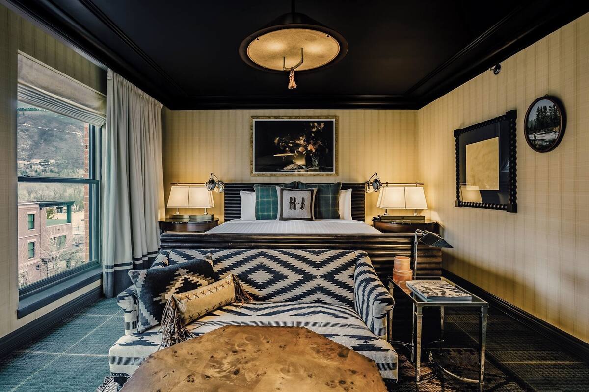 Une chambre d’hôtel avec un grand lit, deux tables de chevet avec des lampes, un canapé à motifs, un pouf en bois et une vue sur le paysage urbain par une grande fenêtre.