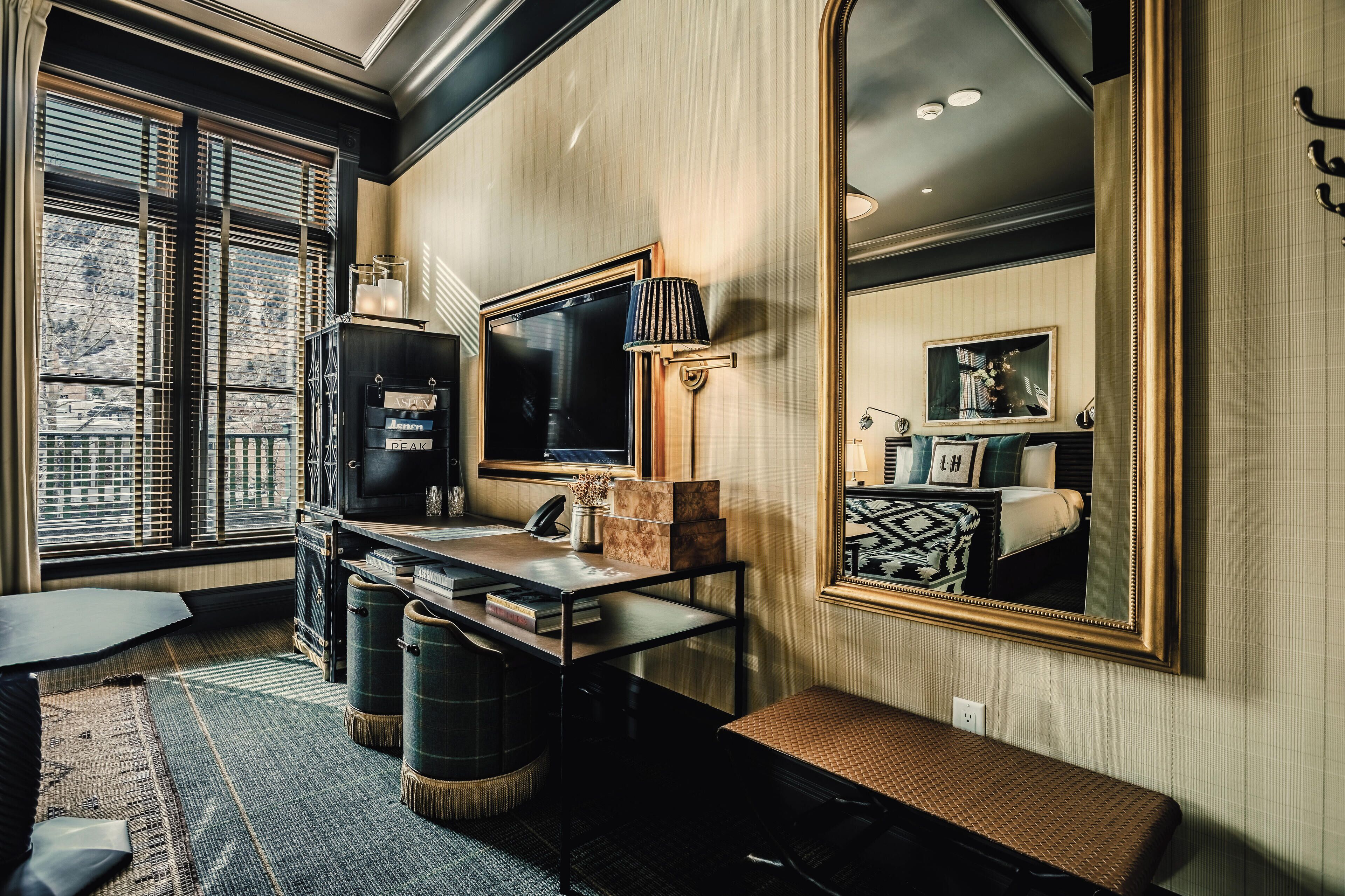 Une pièce avec un grand miroir, un bureau avec une lampe, un téléviseur et une fenêtre avec des stores.