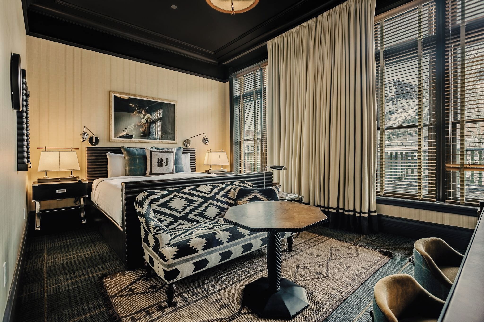 Quarto de hotel com cama, sofá, mesinha, cadeira, luminária, quadro e janela com cortinas.