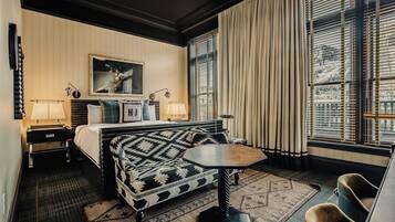 Quarto de hotel com cama, sofá, mesinha, cadeira, luminária, quadro e janela com cortinas.