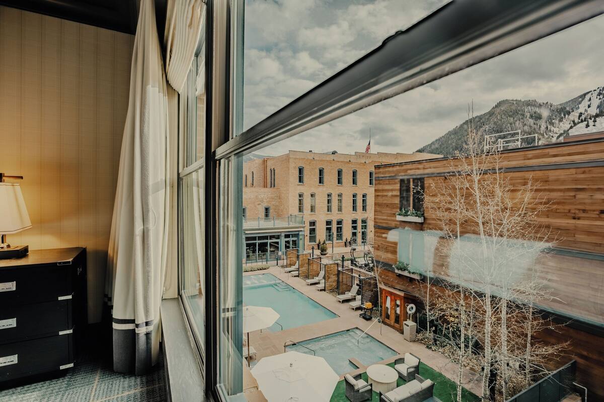 Une chambre d’hôtel dotée d’une grande fenêtre donnant sur un espace piscine, un coin salon extérieur et offrant une vue sur les montagnes.