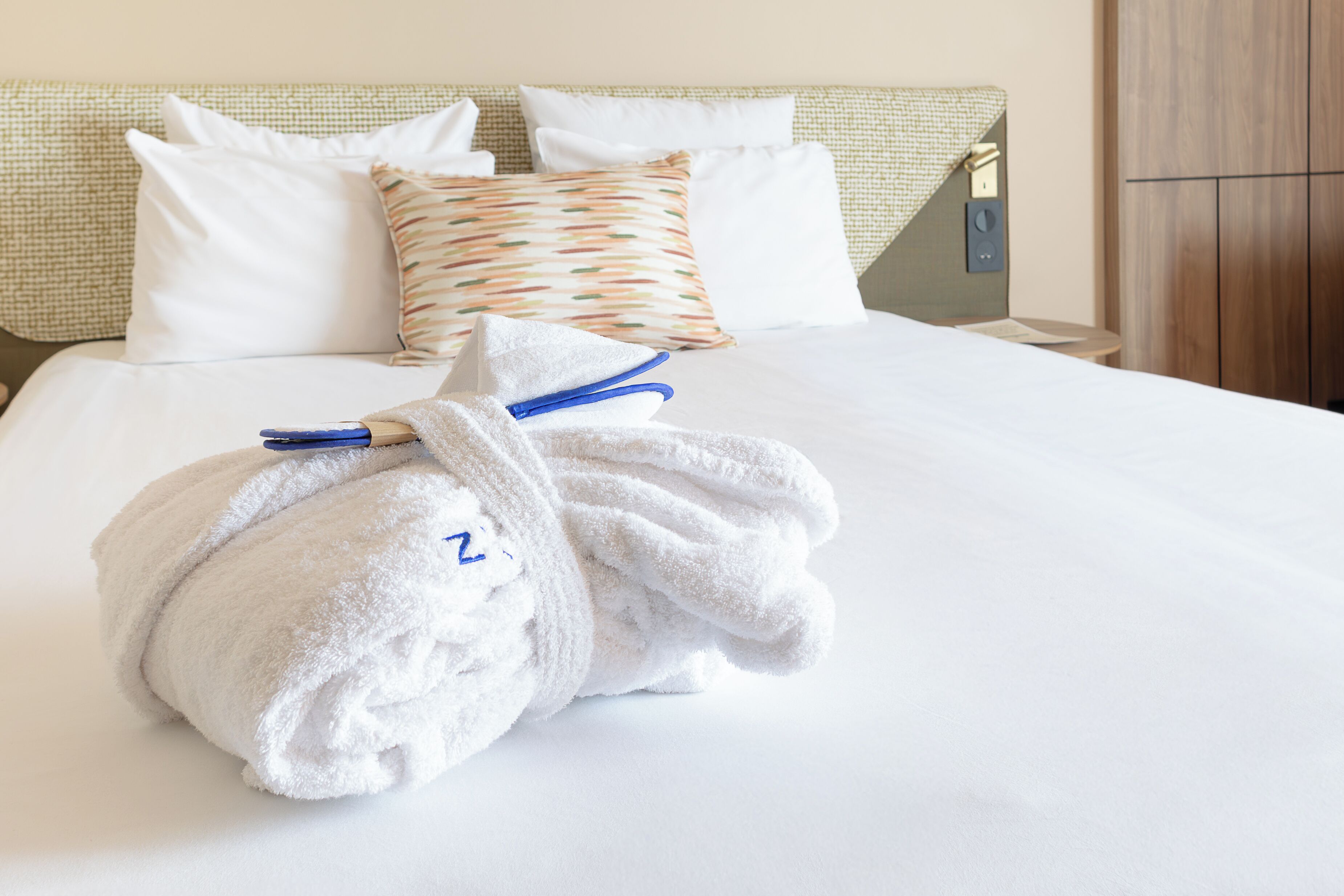 A neatly made bed with white linens, a patterned pillow, and a towel with a blue tag.