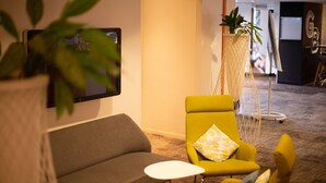 A modern living room with a sofa, a yellow armchair, and a small white table. There is a plant in a white pot and a large potted plant in the background. - Novotel Toulouse Purpan Aéroport (Toulouse)
