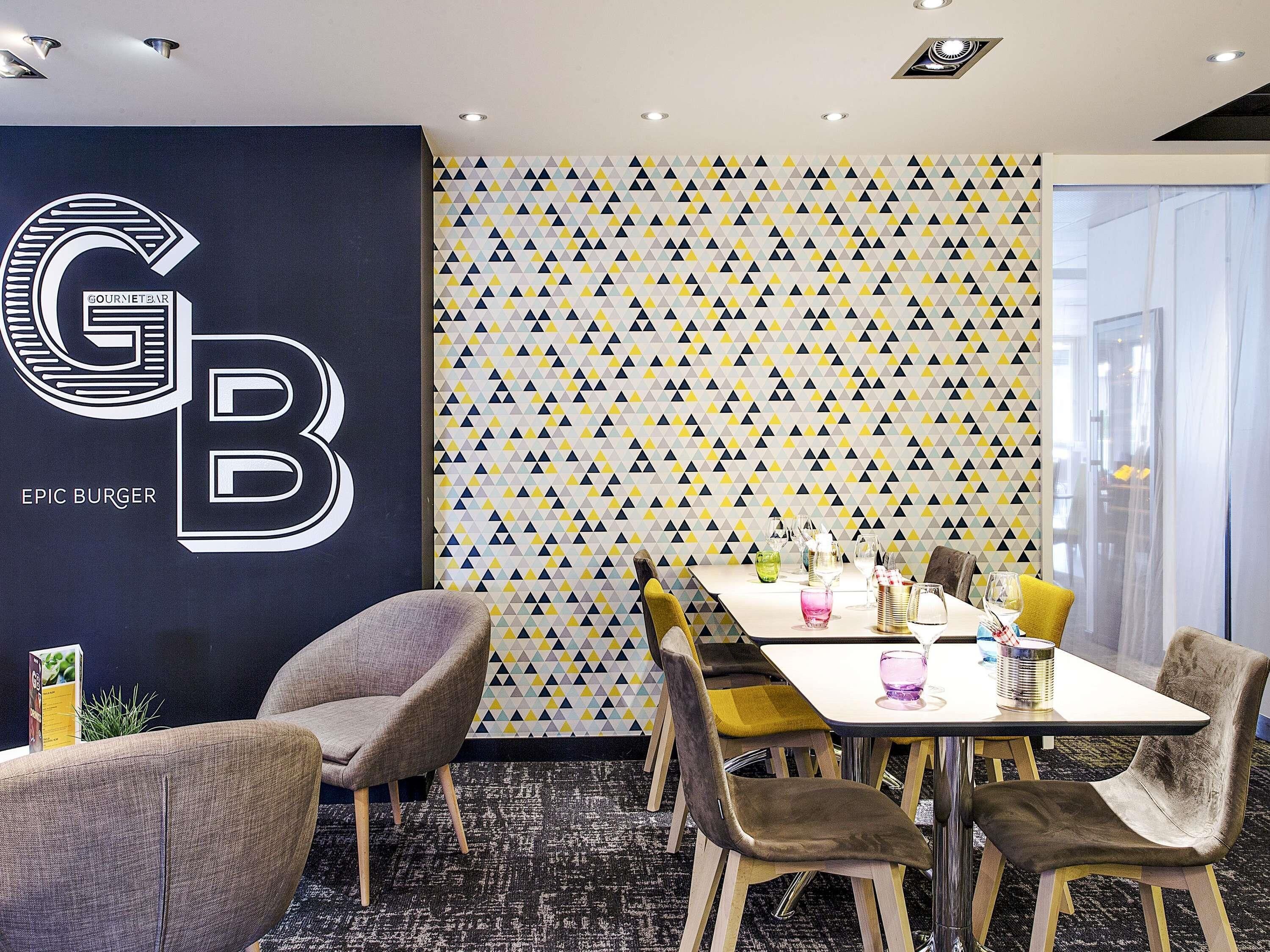 Un espace repas avec un mur orné d’un motif géométrique, un grand logo « GB » et une table entourée de chaises.