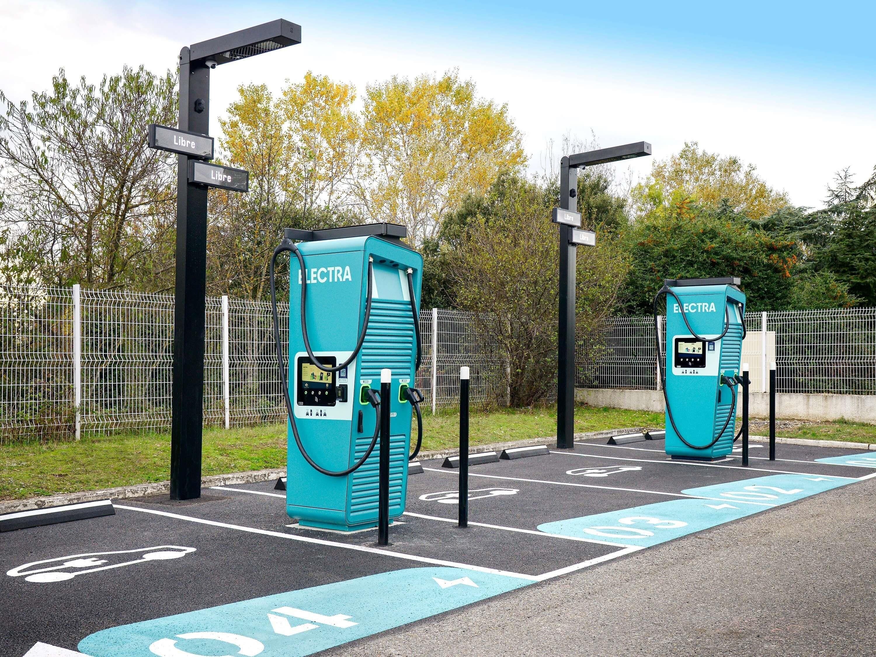 Dos estaciones de carga para vehículos eléctricos, identificadas con el rótulo 'Electra', con un área de estacionamiento cercada y flechas direccionales en el pavimento.