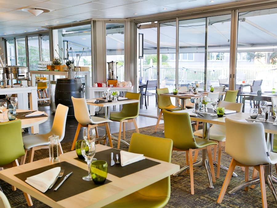 Een modern restaurant met grote ramen, groene en witte stoelen en keurig opgestelde tafels.