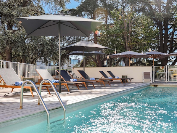 Zona junto a la piscina con sillones de descanso y sombrillas, rodeada de árboles.
