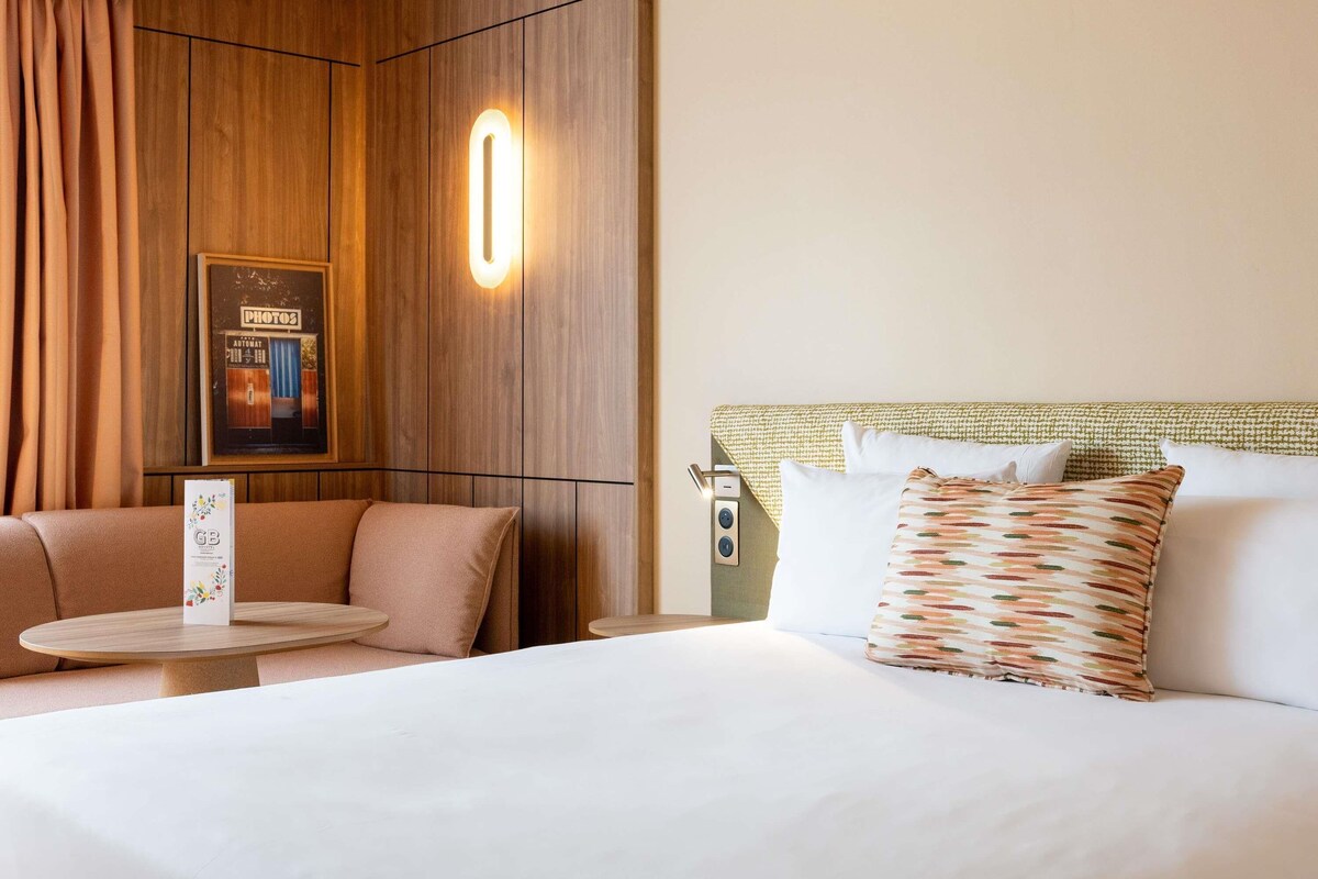 A hotel room with a bed, a small table, a sofa, and a wall-mounted light.