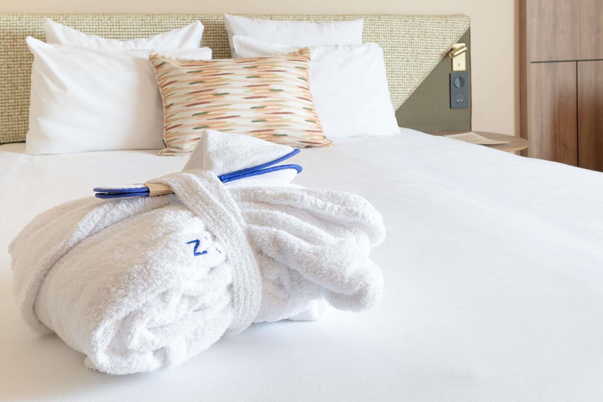 A neatly made bed with white linens, a folded towel with a letter 'Z' on it, and a patterned pillow.