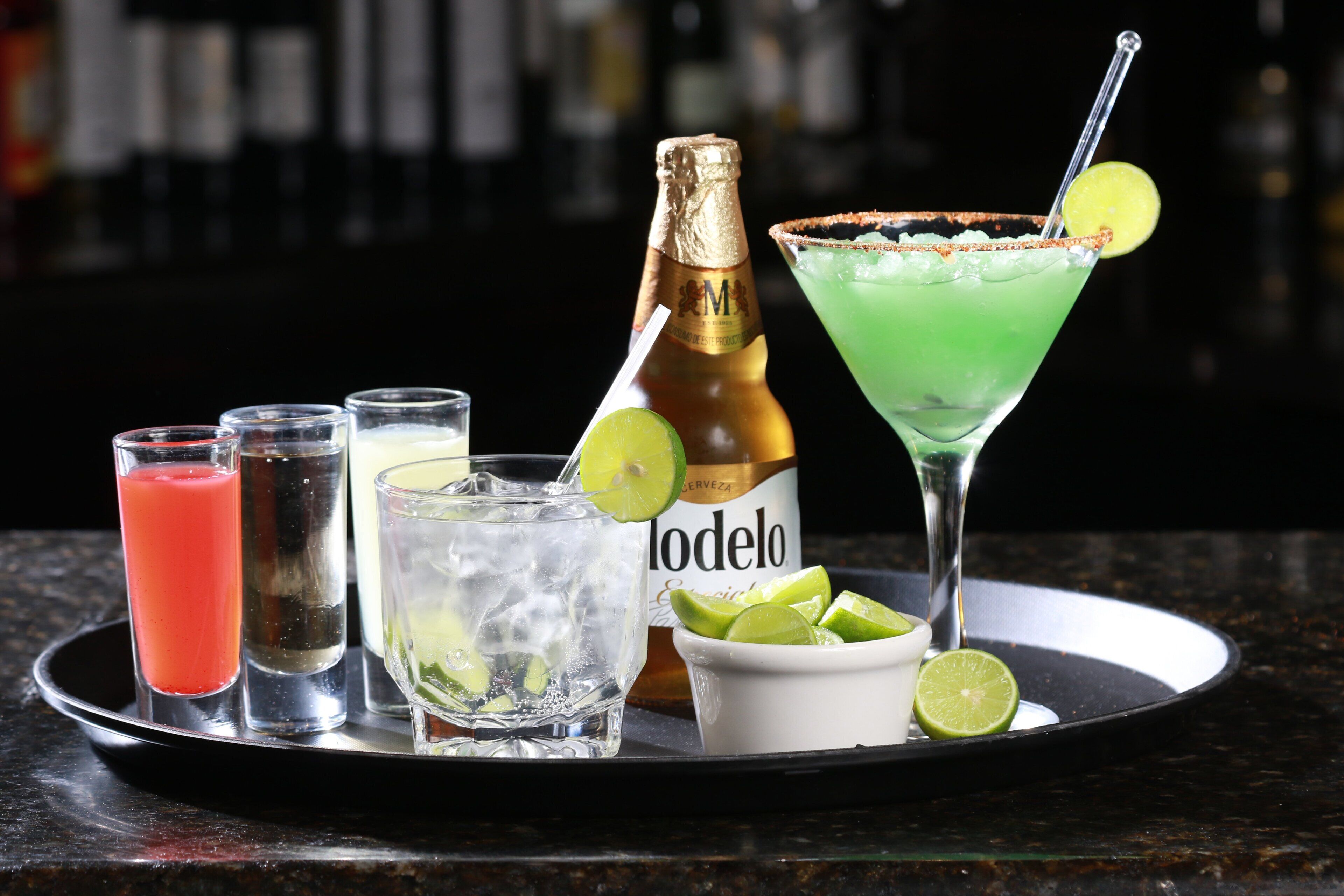 A tray with various drinks, including a margarita, a red cocktail, and a lime-infused beverage, along with a bottle of Modelo beer and a bowl of lime slices.