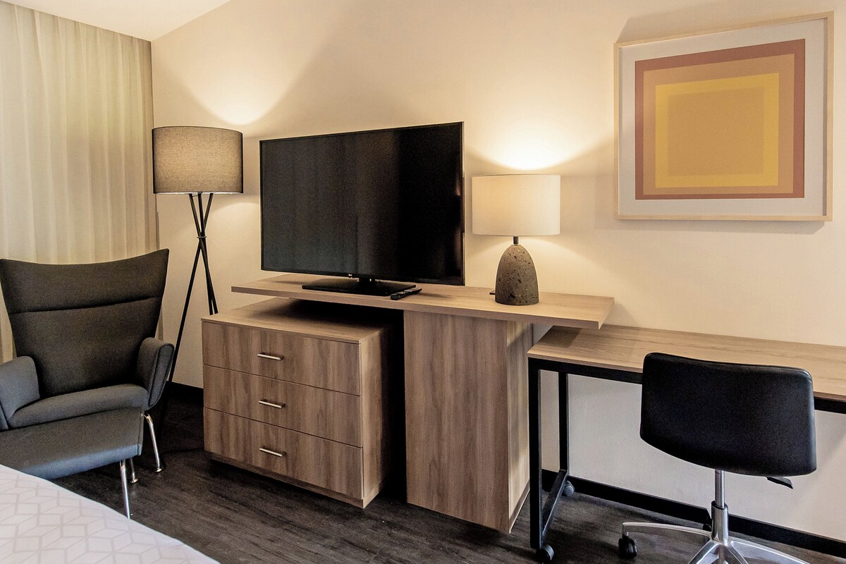 Habitación de hotel con cama, escritorio, televisor de pantalla plana, lámpara, silla y armario.