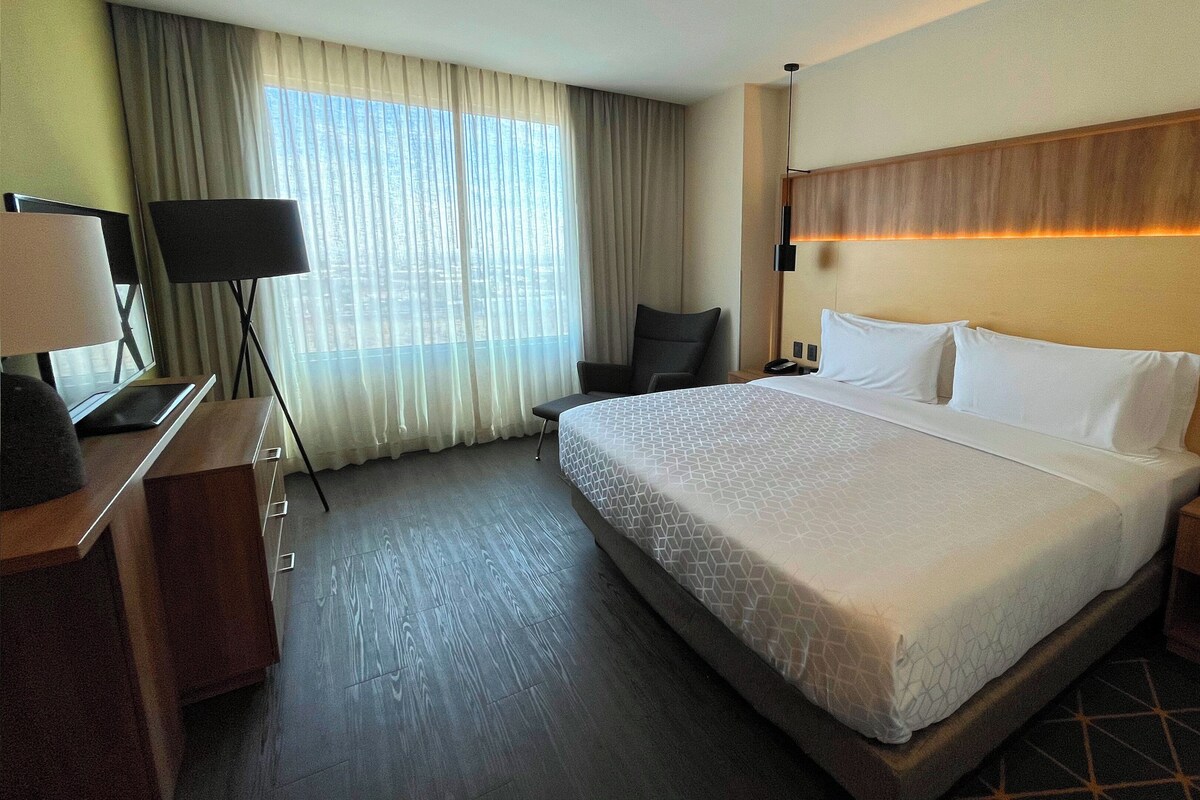Habitación de hotel con una cama grande, un escritorio con lámpara, una silla y vista a la ciudad por la ventana.