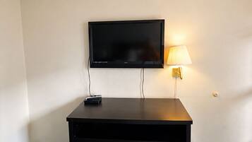 A hotel room with a flat-screen TV mounted on the wall, a desk with drawers, a lamp, and a carpeted floor.