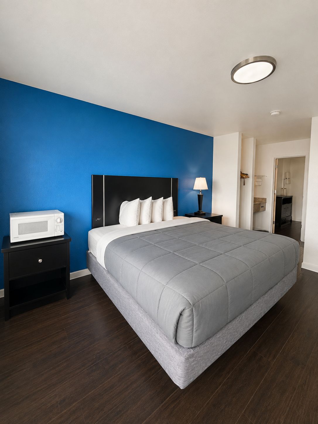 A hotel room with a bed, a nightstand, a microwave, and a door leading to a bathroom.