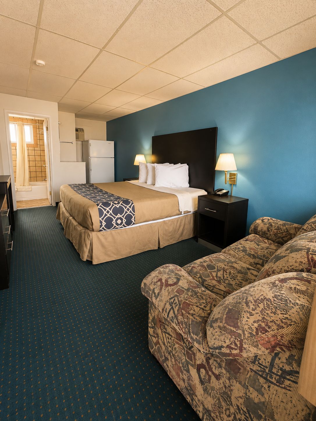 A hotel room with a bed, a sofa, a desk, a lamp, and a refrigerator.