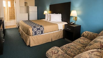 A hotel room with a bed, a sofa, a desk, a lamp, and a refrigerator.