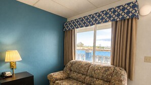 A hotel room with a patterned sofa, a bedside table with a lamp, a window with curtains, and a view of the outdoors. - Rodeway Inn (Vernal)