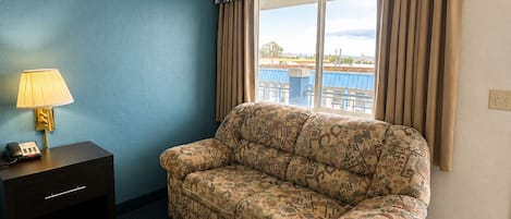 A hotel room with a patterned sofa, a bedside table with a lamp, and a view of the outdoors.