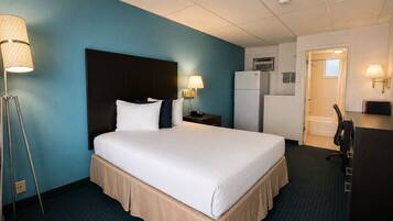 A hotel room with a large bed, a desk, a refrigerator, a lamp, and a bathroom door.