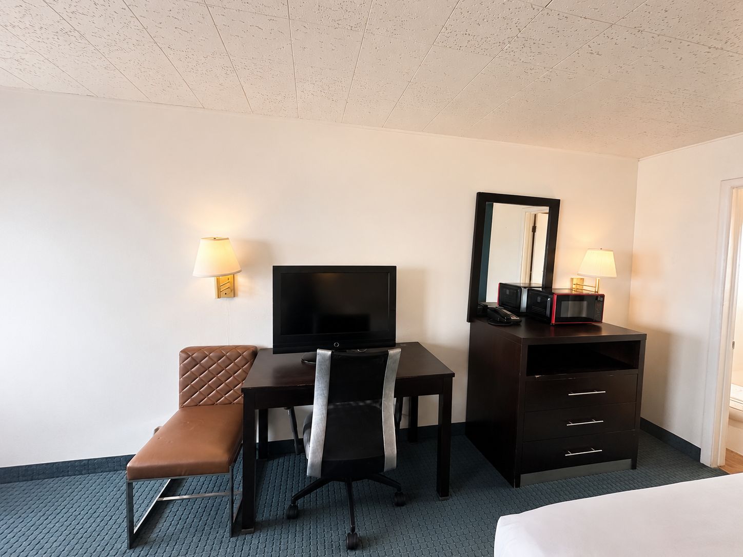 A hotel room with a bed, a desk with a television, a chair, a bench, a dresser, and a mirror.