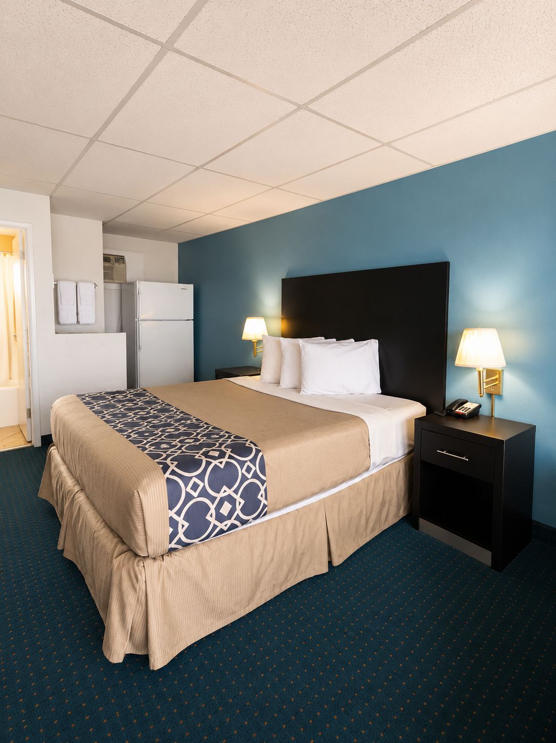 Une chambre d’hôtel avec un lit, deux tables de chevet avec des lampes, un réfrigérateur et une salle de bain visible au fond.