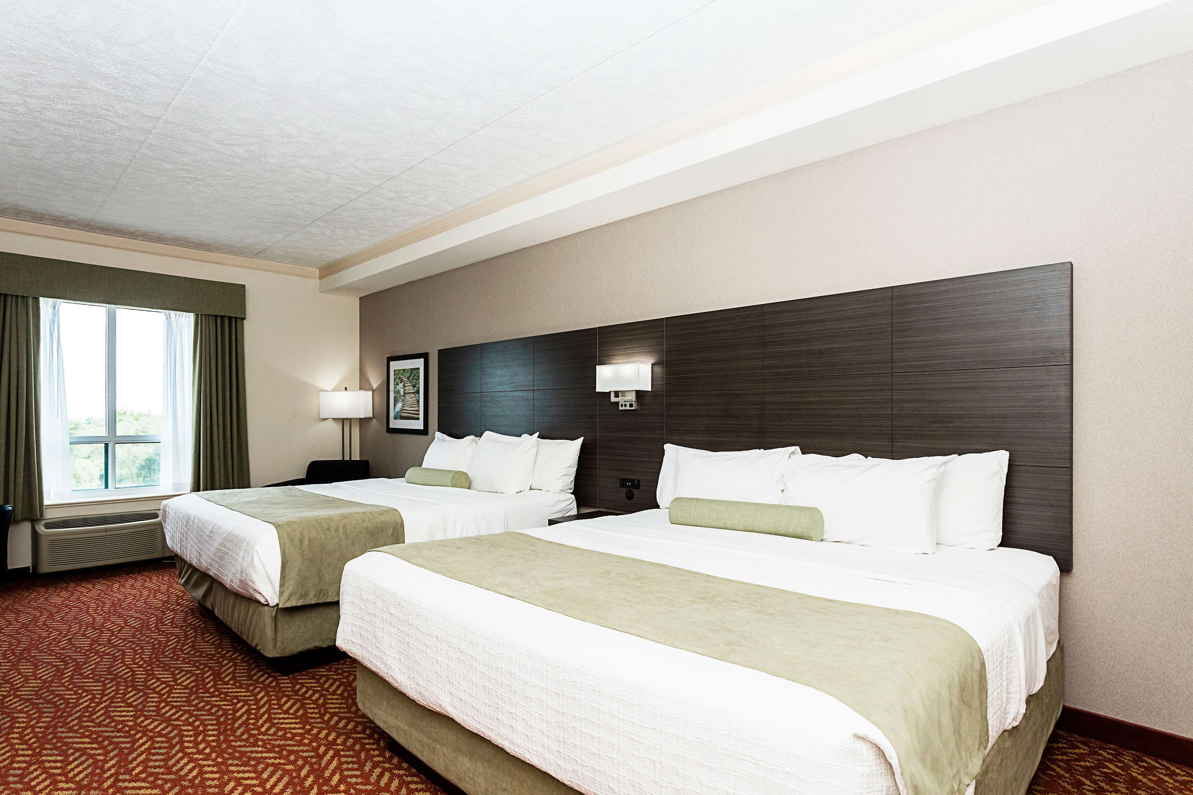 A hotel room with two beds, a large window with curtains, a wall-mounted lamp, and a framed picture.