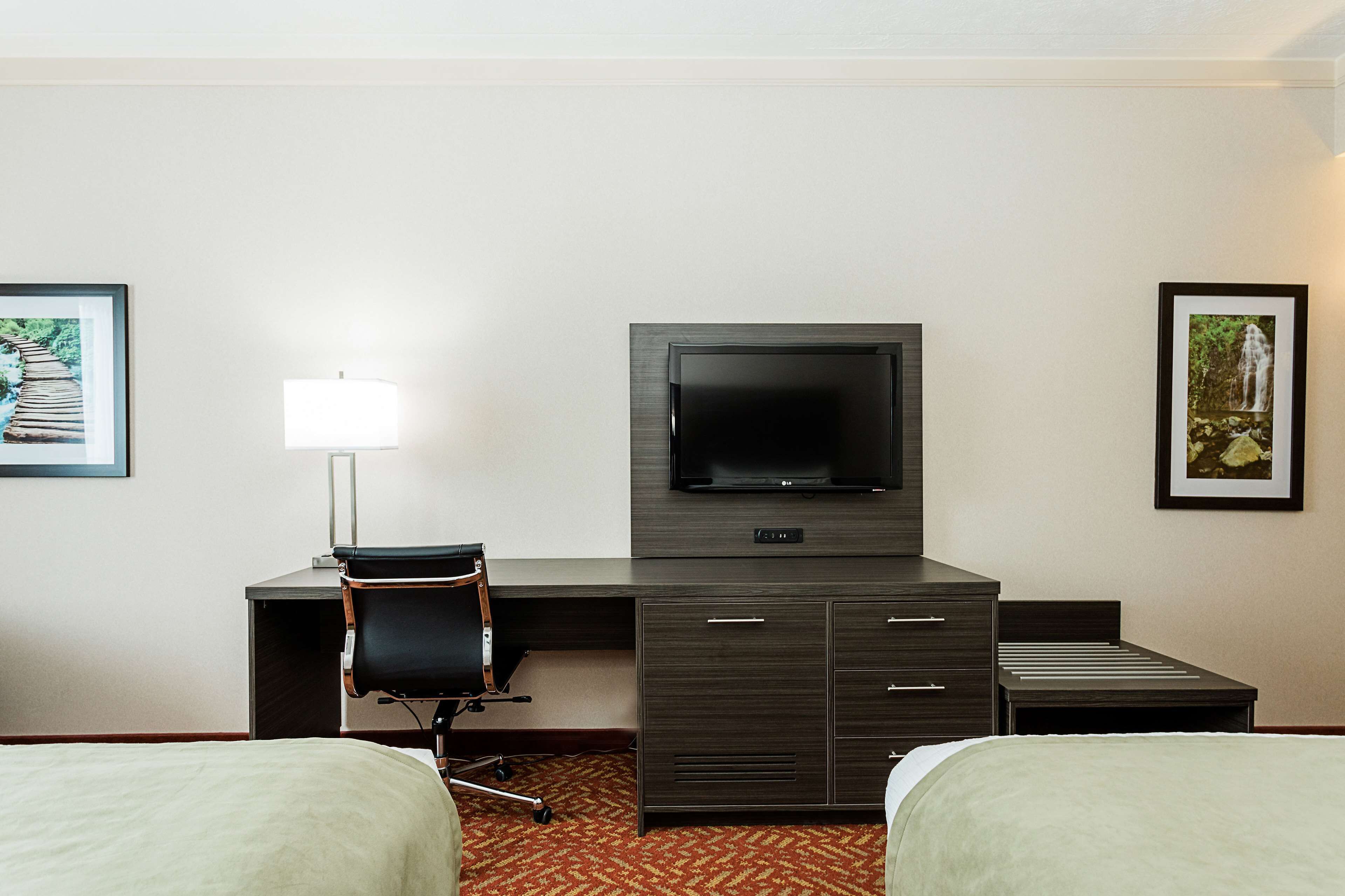 A hotel room with a bed, a desk with a chair, a television, and two framed pictures on the wall.