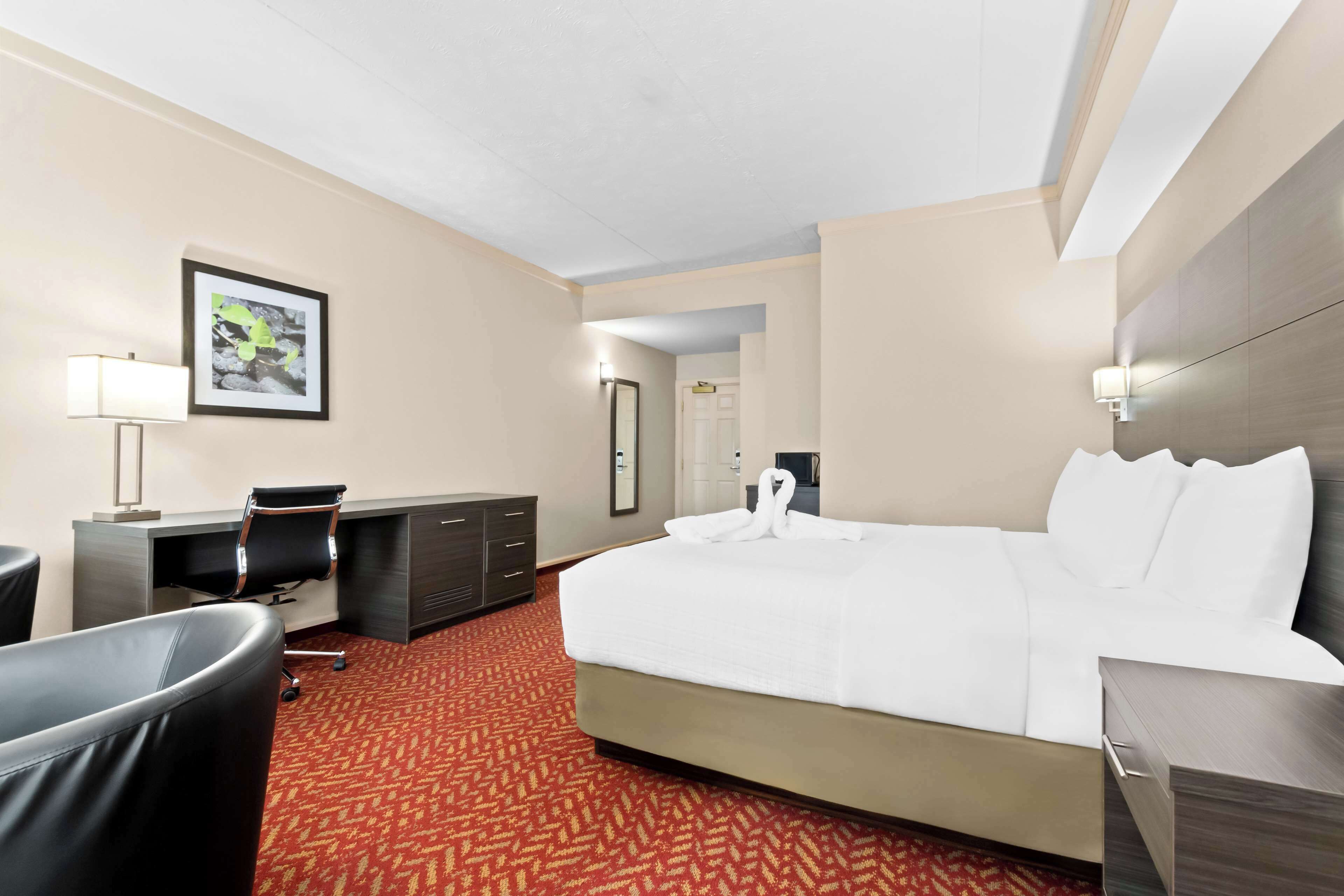 A hotel room with a large bed, a desk with a chair, a desk lamp, a mirror, and a framed picture on the wall.