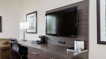 Habitación de hotel con una televisión de pantalla plana montada en la pared, un escritorio con lámpara, una silla y una cafetera.