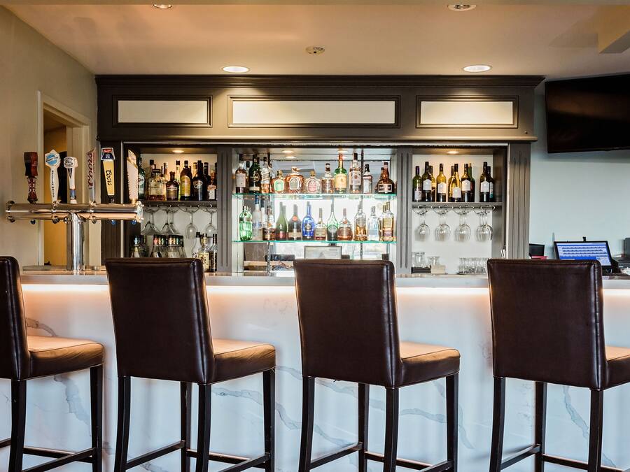 A bar area with a marble countertop, three leather bar stools, a variety of liquor bottles, and a television mounted on the wall.