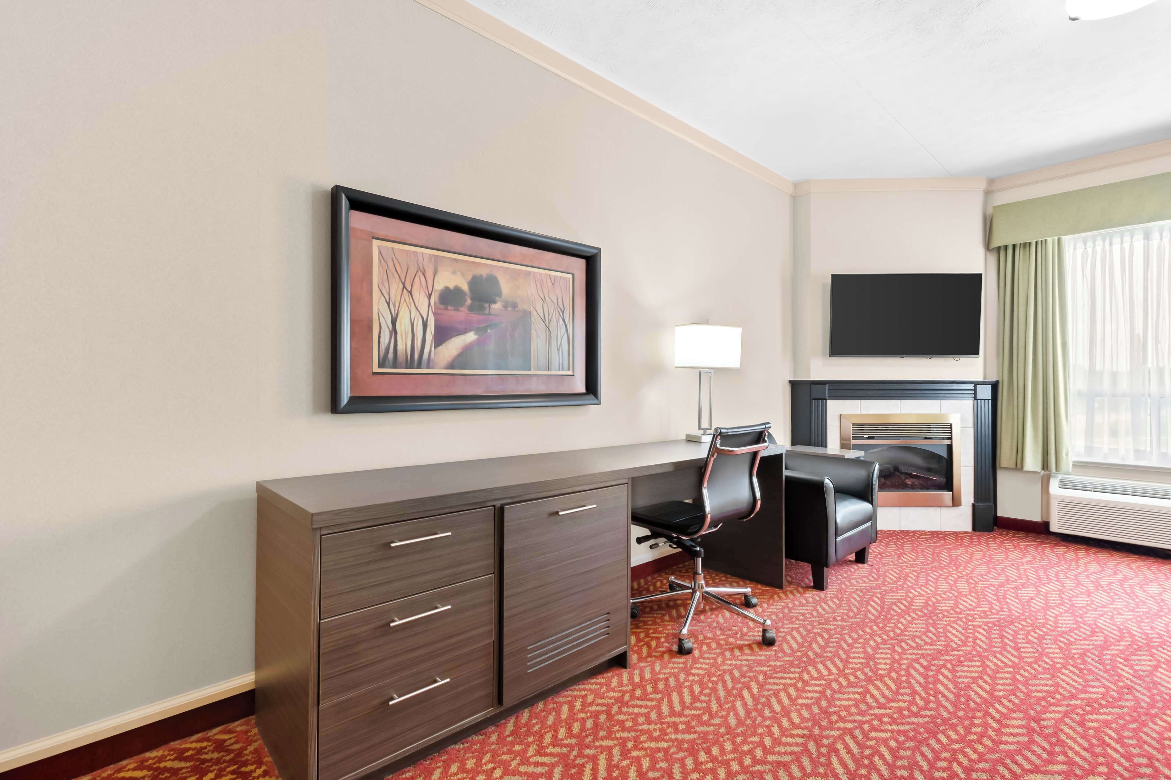 A hotel room with a fireplace, a desk with drawers, a chair, a television on the wall, and a patterned carpet.
