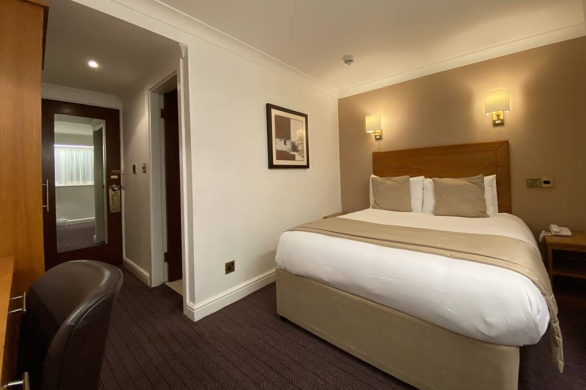 A hotel room with a large bed, a bedside table with a lamp, a chair, and a door leading to another room.