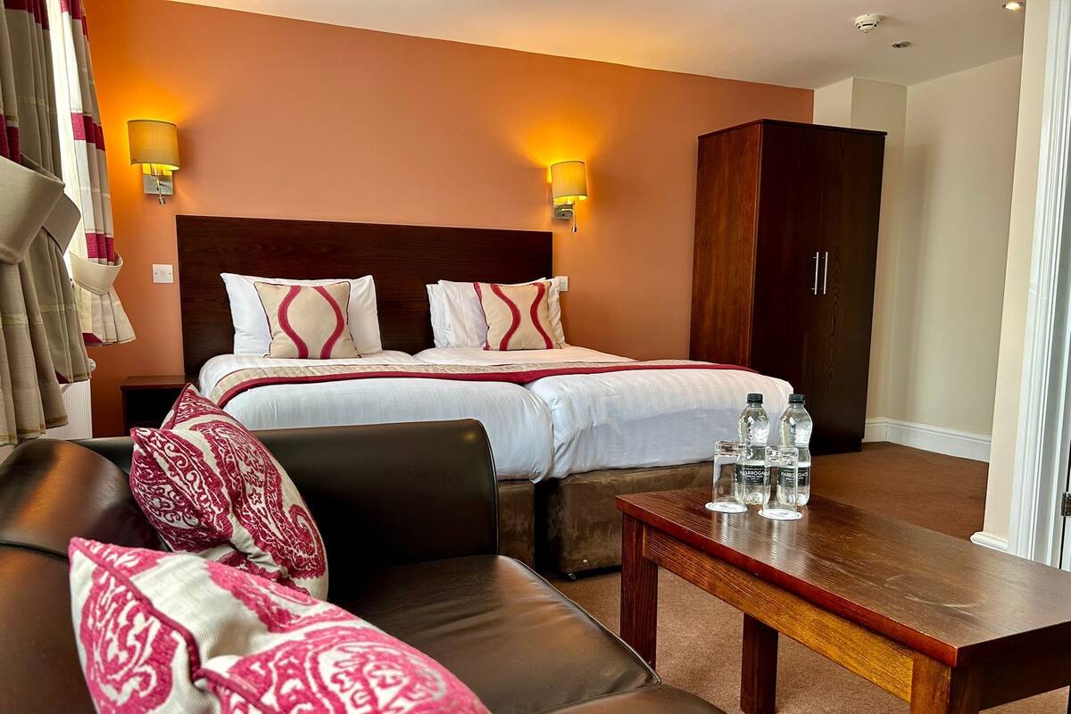 A hotel room with a large bed, a wooden wardrobe, a leather sofa, and a small table with water bottles.