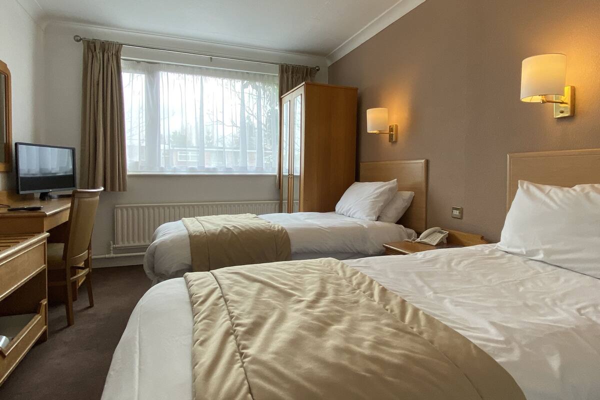 A hotel room with two beds, a desk with a computer, a chair, a window with curtains, and a wall-mounted lamp.
