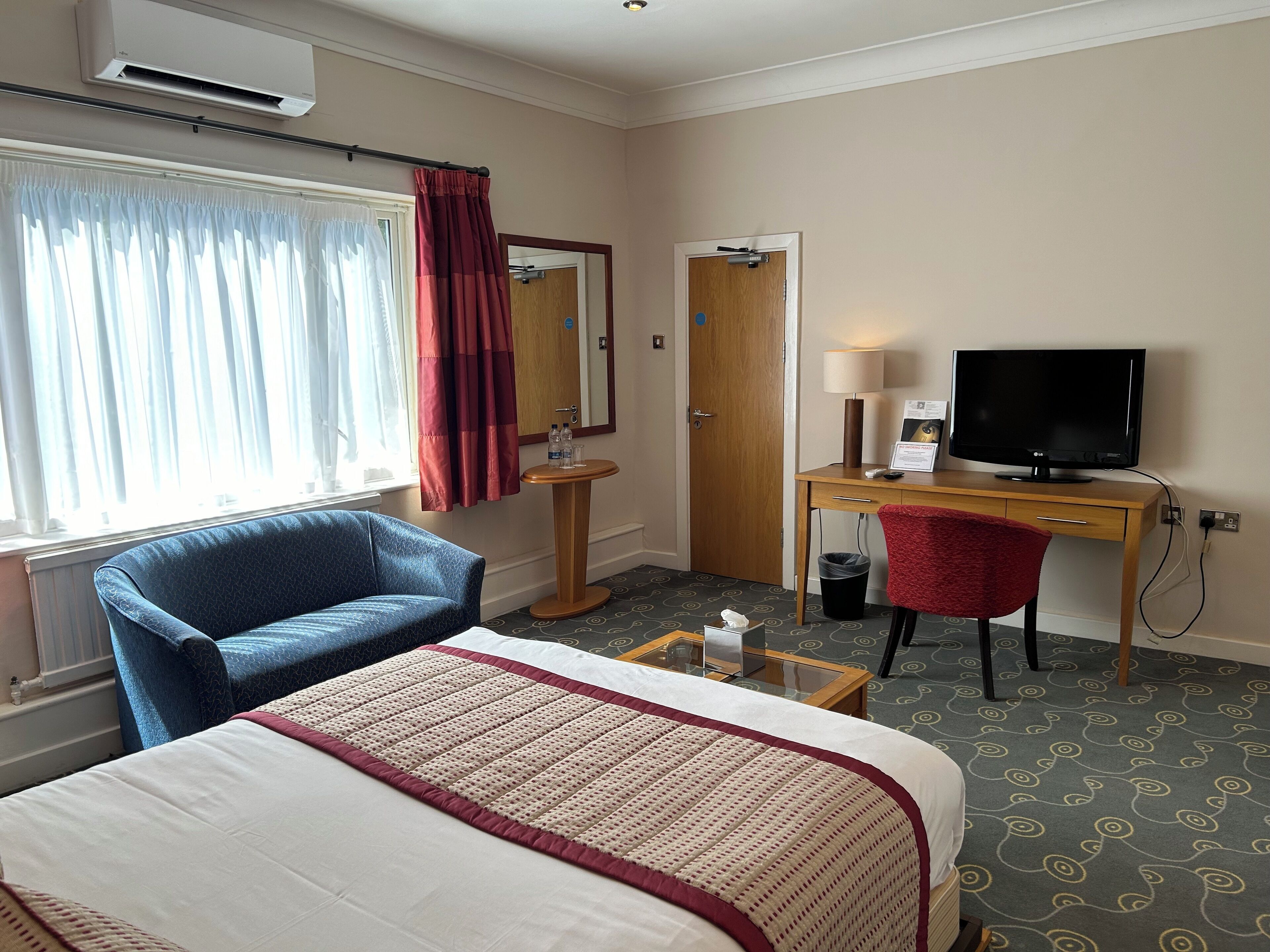 Habitación de hotel con cama, escritorio con computadora, silla, televisor y ventana con cortinas.