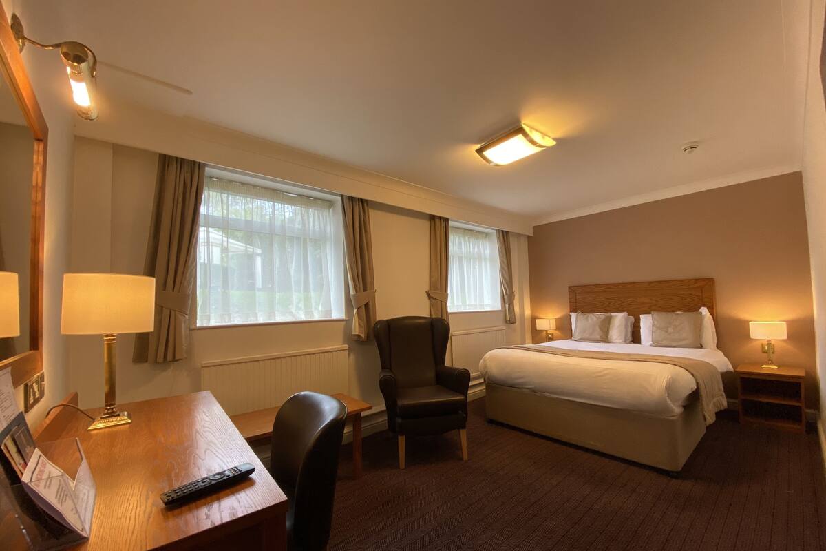 A hotel room with a bed, a desk with a lamp, a chair, and a window with curtains.