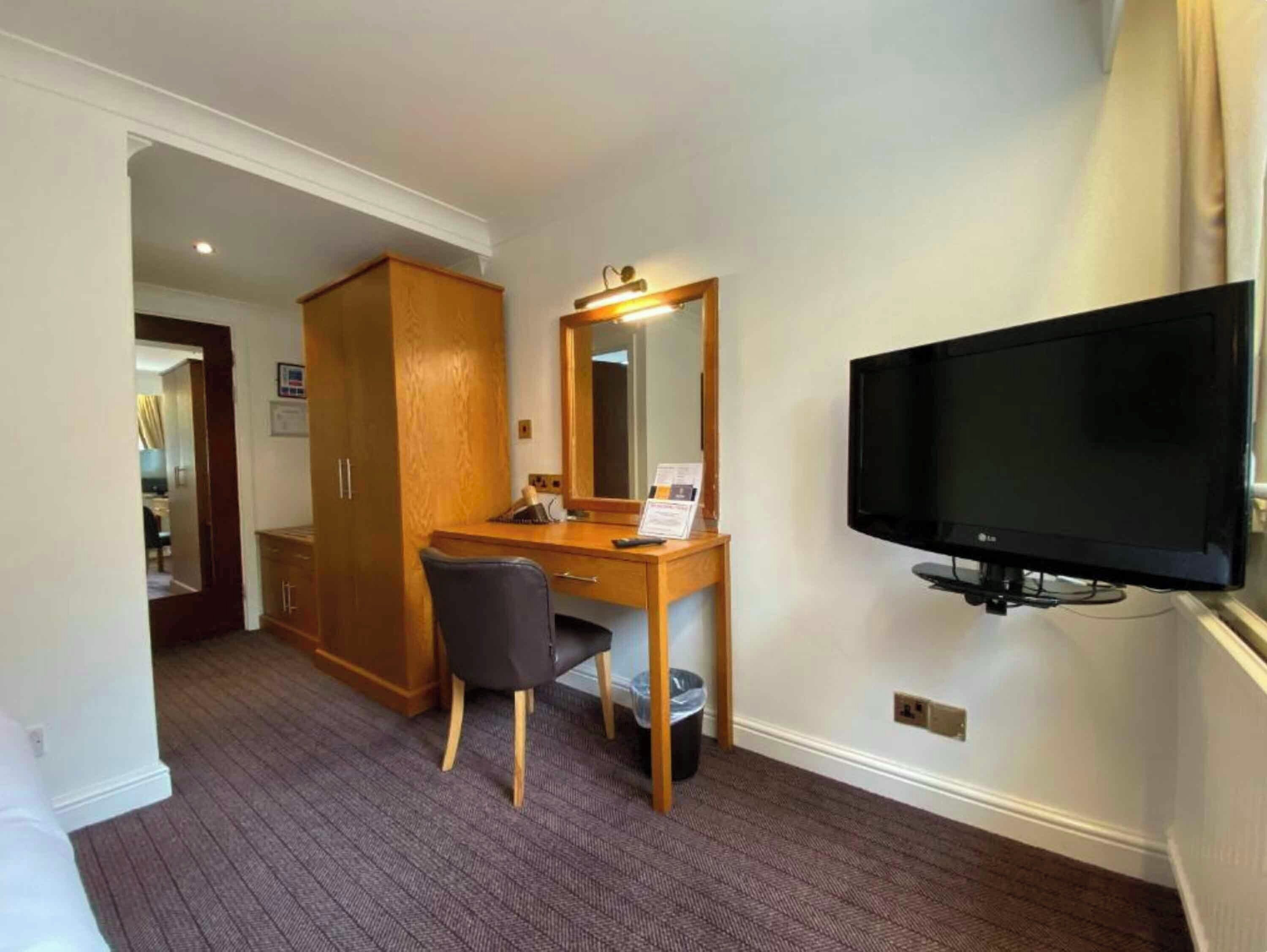 A hotel room with a wooden desk, a chair, a television mounted on the wall, and a wardrobe.