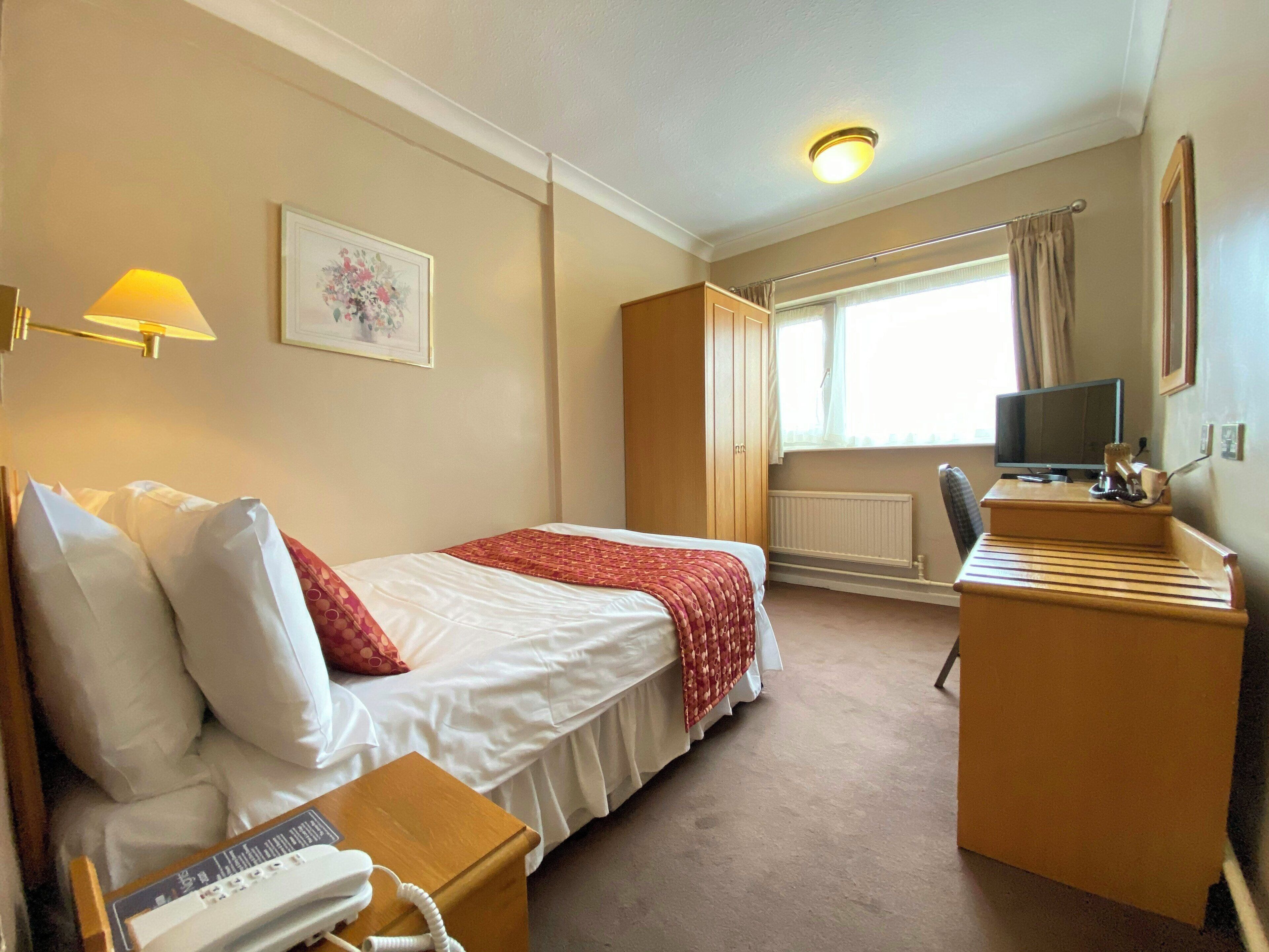 A hotel room with a bed, bedside table, lamp, desk, chair, television, and a window with curtains.