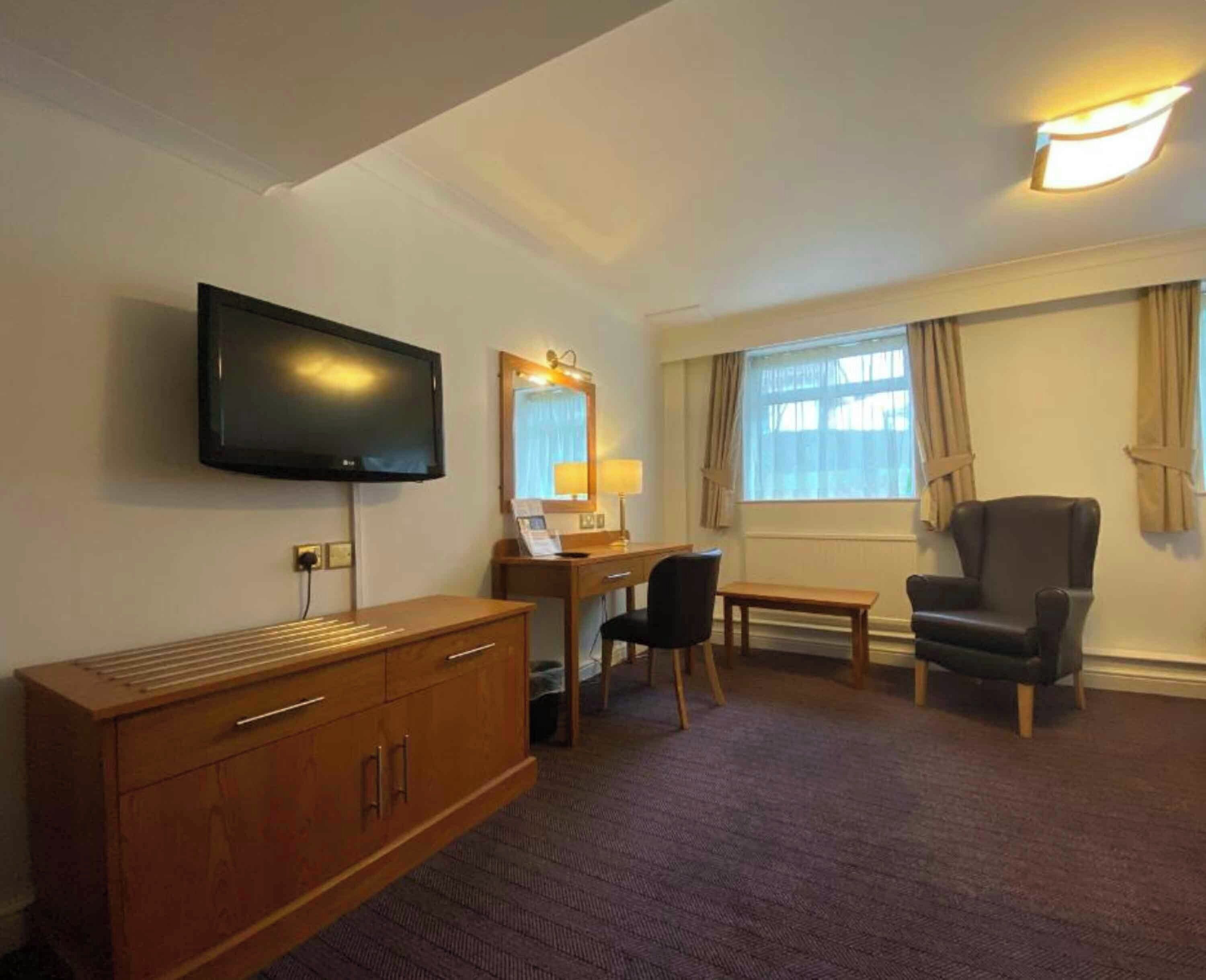 A hotel room with a flat-screen TV, a desk with a chair, a leather armchair, and a large window with curtains.