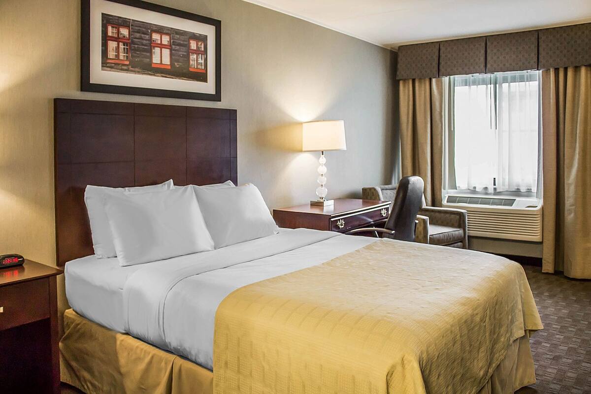 A hotel room with a large bed, a desk with a lamp, a chair, a window with curtains, and a framed picture on the wall.